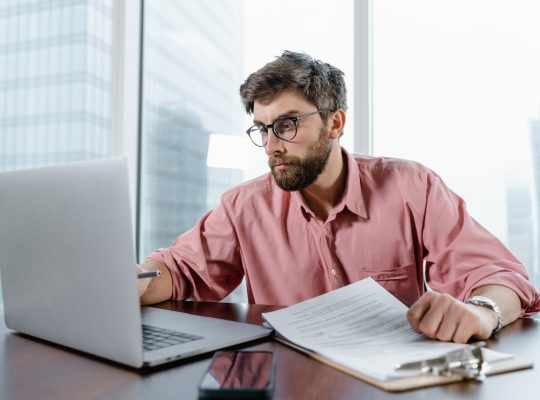 A businessman in an office reviews documents and data on a laptop, concentrating on financial analysis.