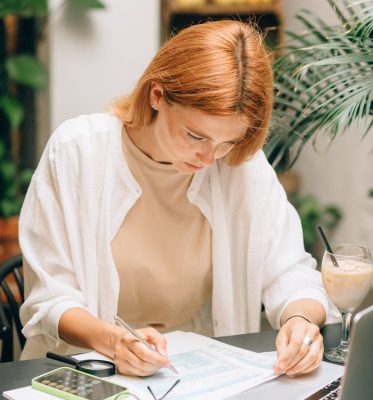Woman analyzing financial documents using laptop and calculator indoors.