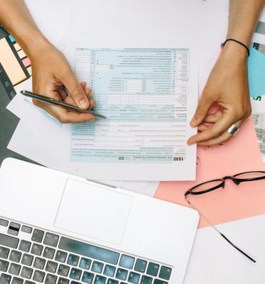 Hands writing on tax documents with laptop, glasses, and currency on desk.